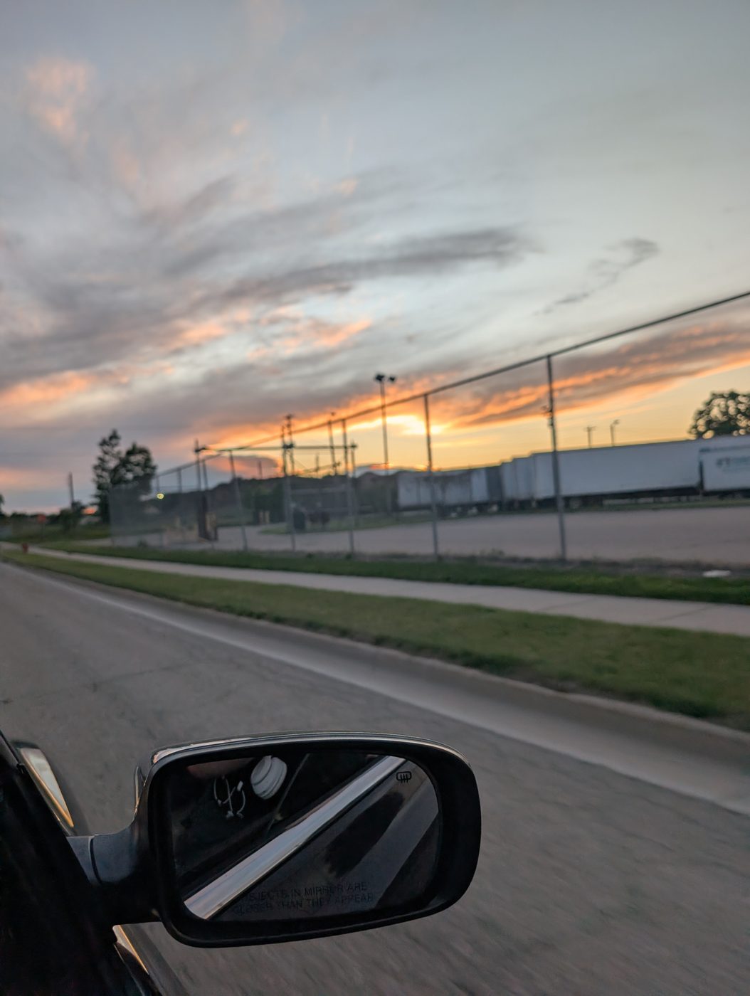 A sunset in orange and pink over a chain-link ballfield, parking lot, and semi-trailers, seen past a car's side mirror
