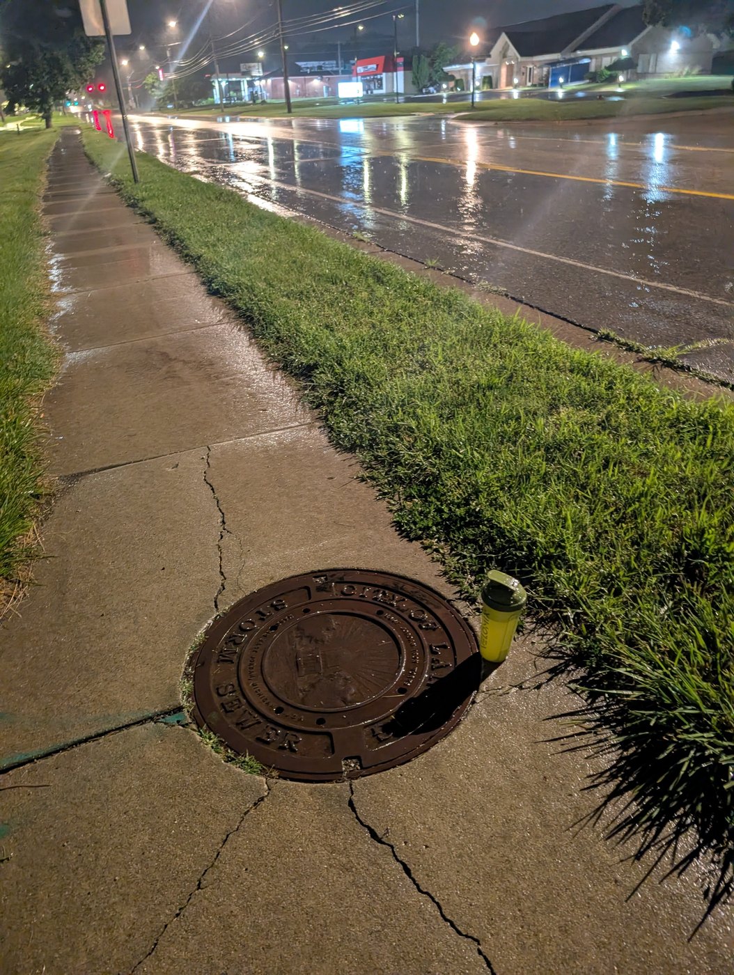 A cast-iron sewer cover on a wet, cracked sidewalk at night, a rain-slicked street with streetlights and storefronts reflected behind it, and a small yellow bottle resting at the edge