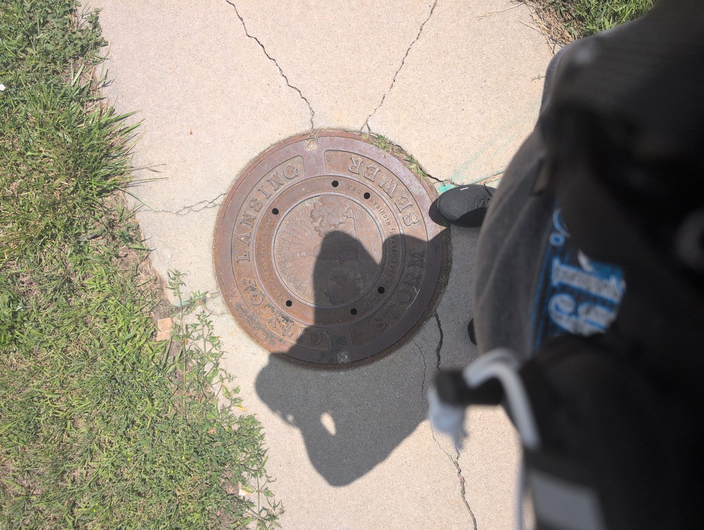 A rusted City of Lansing cast-iron sewer cover on a cracked sidewalk, the photographer's shadow falling across it